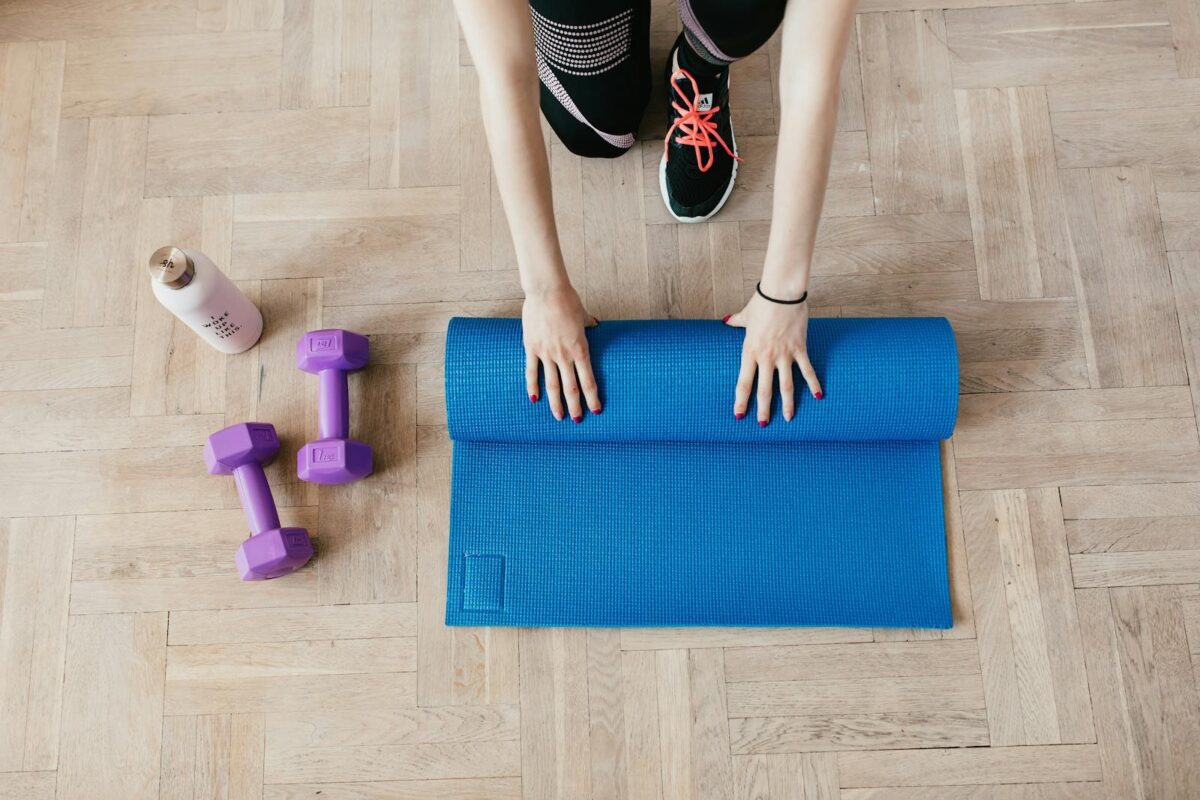 Yogamatte wird auf Parkettboden aufgerollt, daneben Hanteln und Wasserflasche